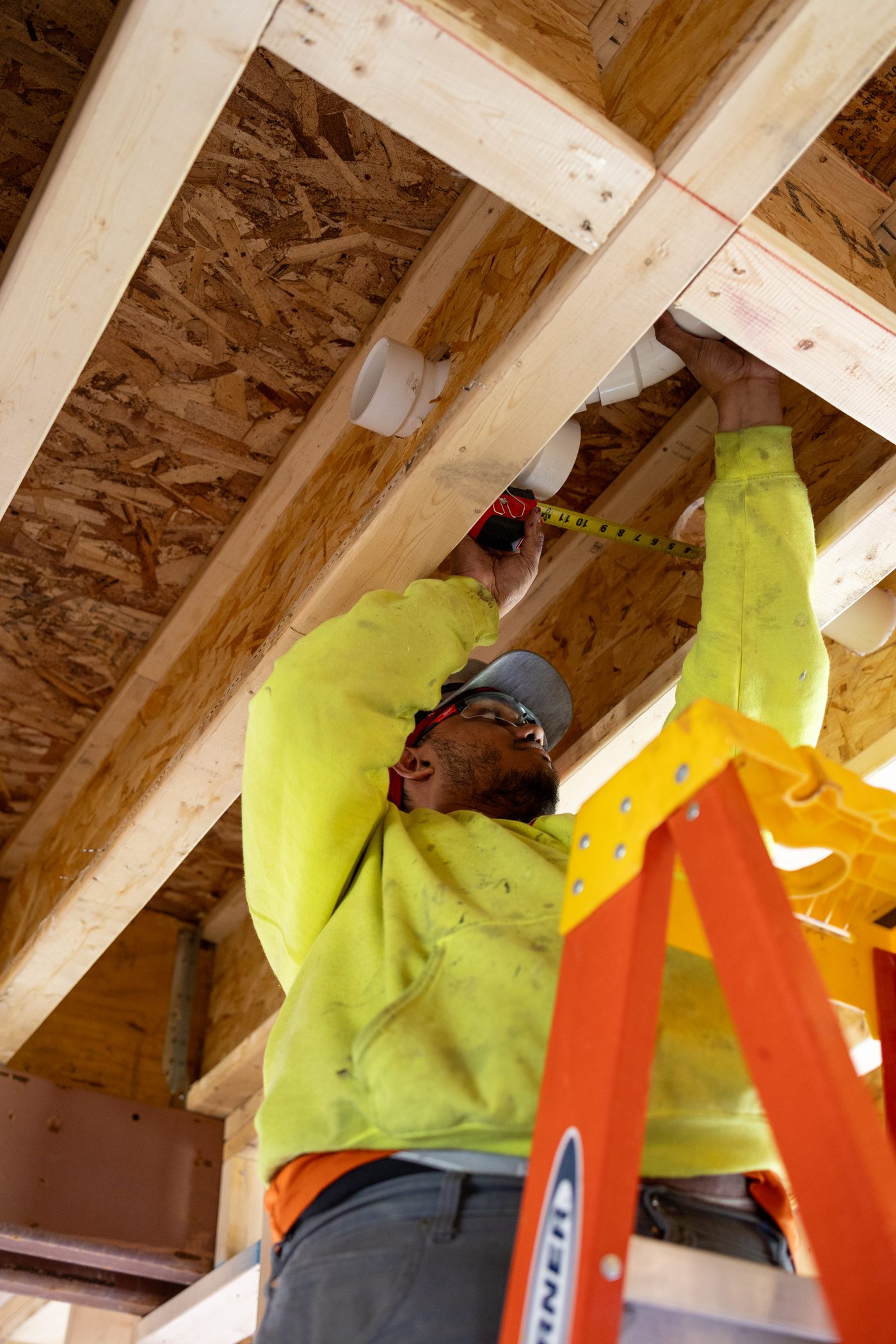 A Hertel plumber measuring an overhead drain rough-in between joists